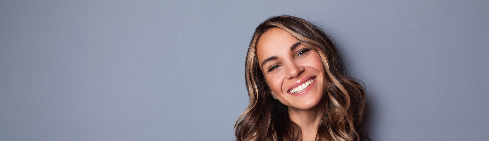 The image is a photograph of a woman with light skin, smiling at the camera. She appears to be in her late twenties or early thirties and has long hair. Her eyes are looking directly at the camera, and she is holding up her index finger near her mouth as if she s making a point or emphasizing something. The background is plain and light-colored, which suggests that this could be a stock photo used for various purposes such as advertising, personal branding, or lifestyle content.
