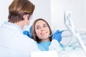 Smiling young woman is sitting in dental blue chair in clinic, office. Man doctor, orthodontist is conducting examination, treating teeth with tools, instruments. Visit to dentist concept.