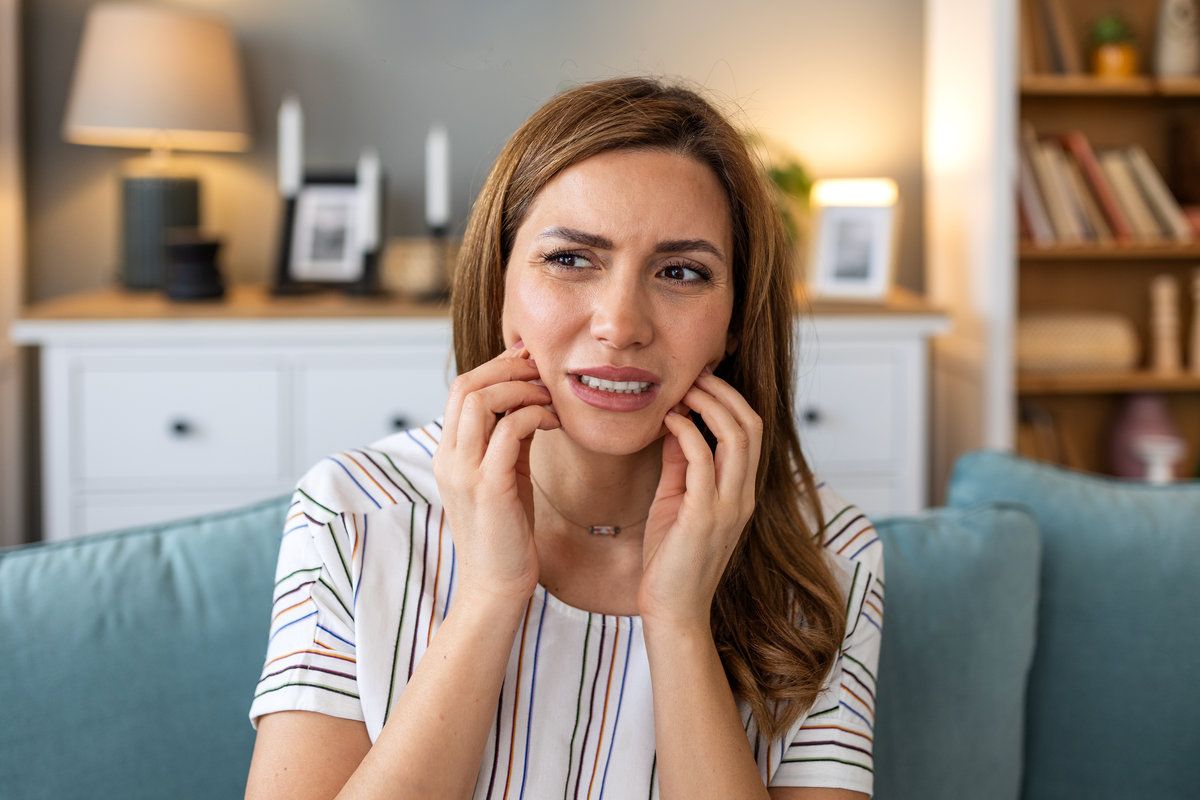 Toothache and dental pain. Attractive woman sitting on sofa holding painful jaw and cheeks with hands, Healthcare And Medicine concept. Person with teeth problem feeling sensitivity and facial